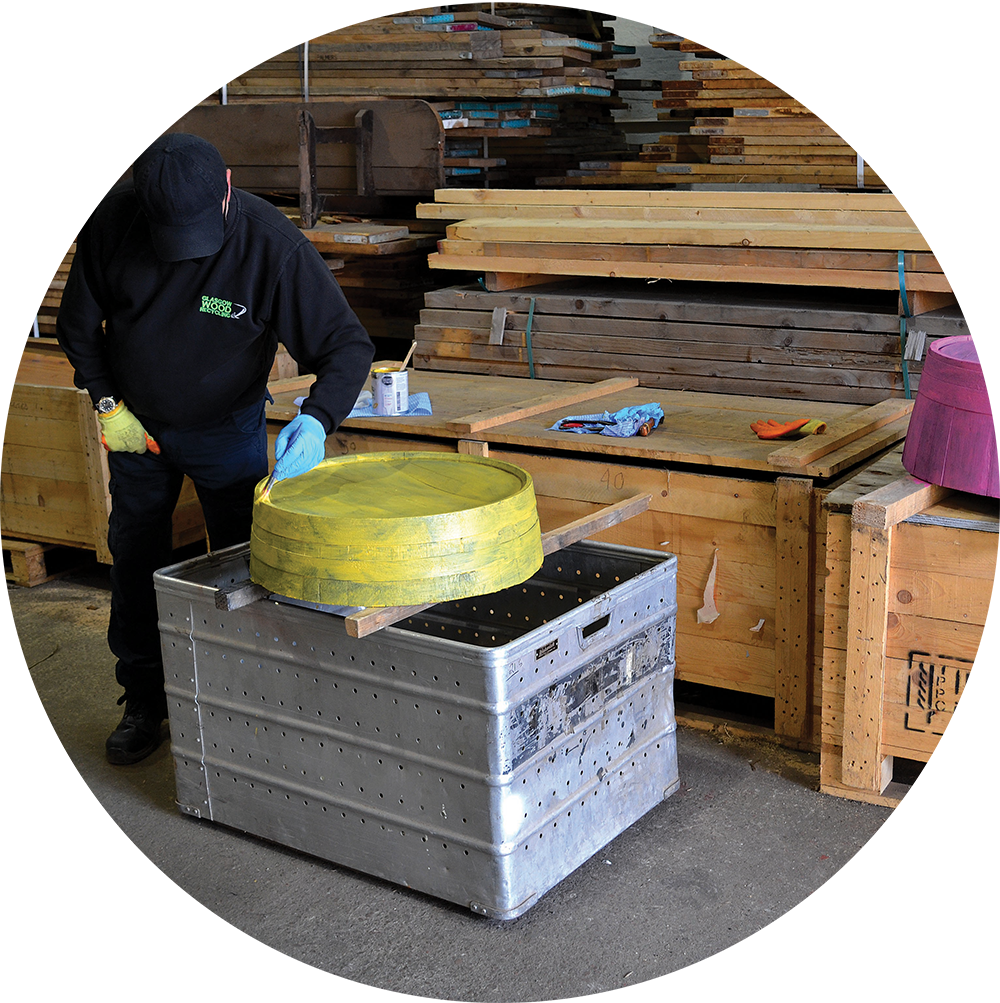 Man standing over wooden cask barrel top section, painted yellow, on top of a silver metal box, surrounded by wooden creates in the background