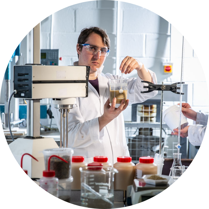 Man in white lad coat, wearing protective eye wear, holding a beaker and string contents with glass tube.  Standing in a lab, surrounded by scientific equipment and materials